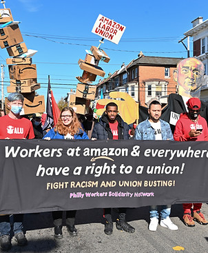 Image shows a multi-racial group of people holding a black banner that reads 'Workers at Amazon & everywhere have a right to a union! Fight racism and union busting!'. Behind the people holding the banner are towers of amazon delivery boxes with a sign at the top that says 'Amazon Labor Union', they are at the Peoplehood Parade in West Philly. Photo credit: Joe Piette