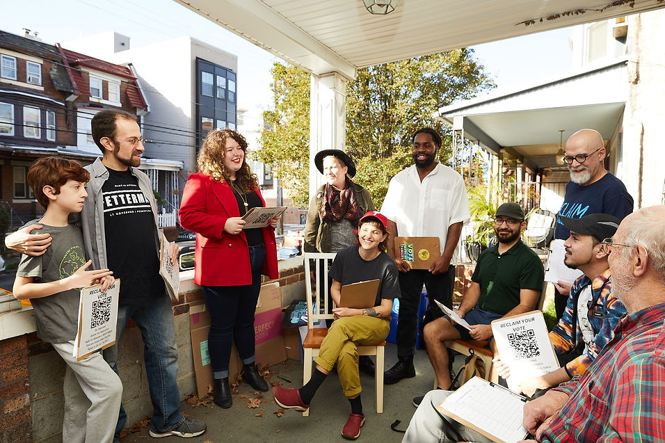 Image shows Amanda, a white woman with curly hair, wearing a red blazer talking on a porch with a mutli-racial, multi-generational group of neighbors sitting and standing in a semi-circle with clipboards ready to go canvassing. Photo credit: Hibbard Nash