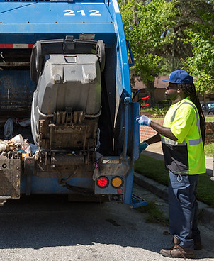 Image: An African American man in a yellow high visibility vest operates a blue trash truck. Photo credit: Ryan Johnson